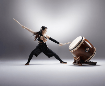 A woman dressed in black martial arts attire performs a dynamic pose with drumsticks in each hand, striking a large taiko drum in Central Oregon.