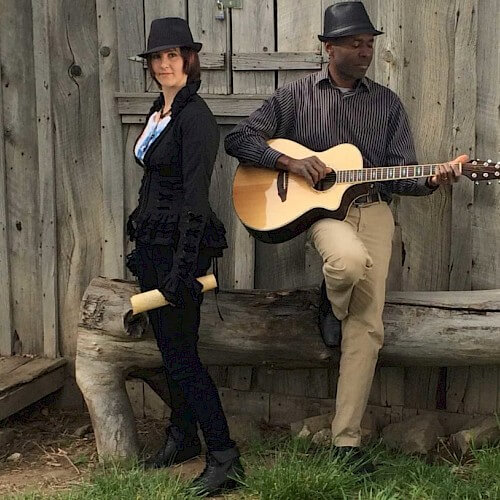 A man and woman dressed in black, with matching hats, sit on a wooden bench, one playing an acoustic guitar in Central Oregon.