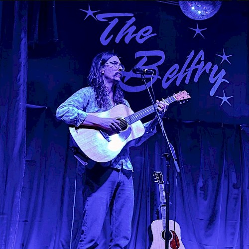 A musician with long hair plays an acoustic guitar on stage at The Belfry in Central Oregon.