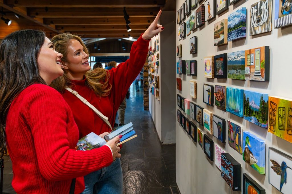 Two women in red sweaters look at small paintings displayed on a gallery wall in Central Oregon.