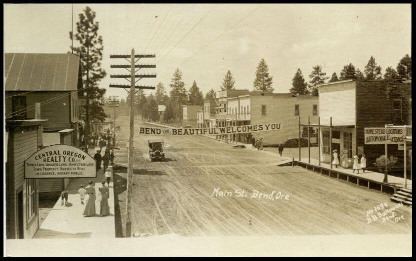 A vintage photograph of a dirt street in Bend, Oregon with a welcome banner in Central Oregon.