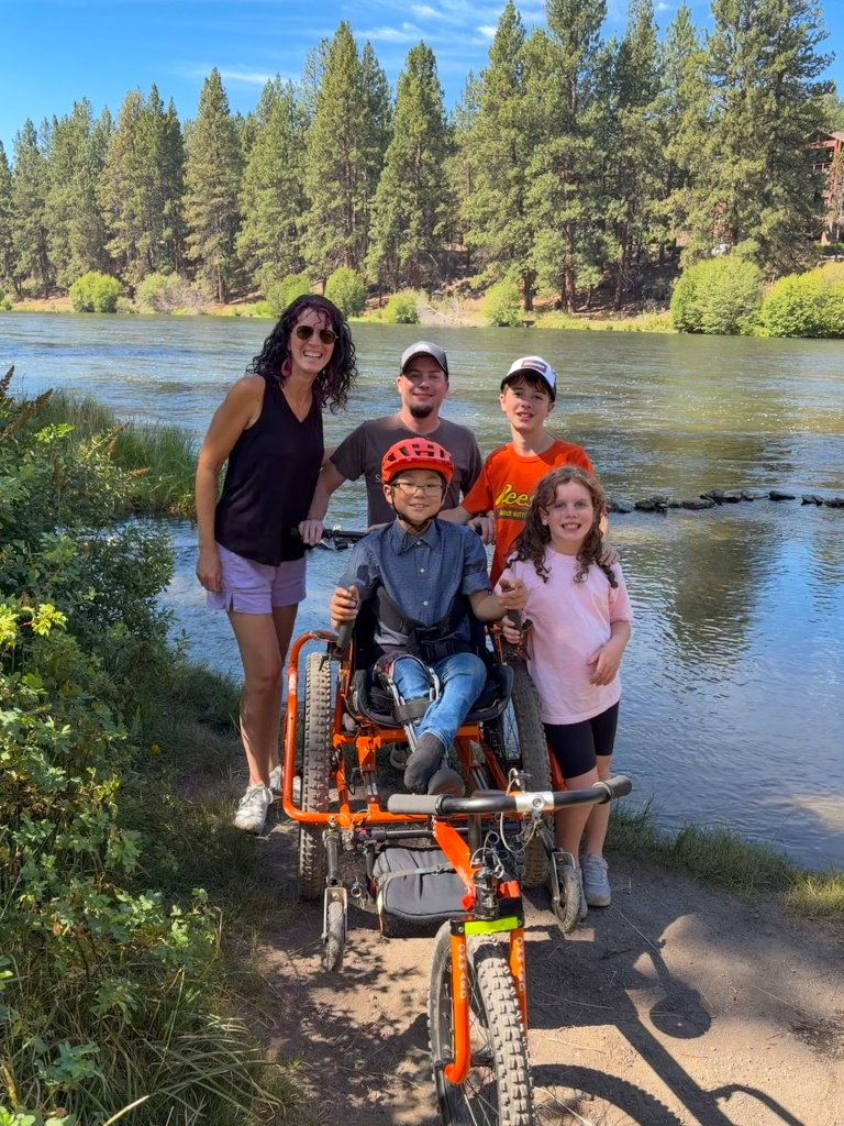 A family poses together by a river with a child in an adaptive bicycle in Central Oregon.