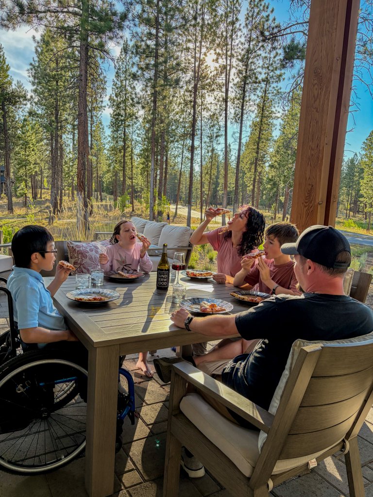 A family eating pizza together on an outdoor patio surrounded by pine trees in Central Oregon.