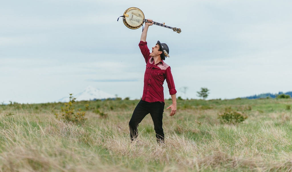 A man in a hat and red shirt holds a banjo high in a field in Central Oregon.