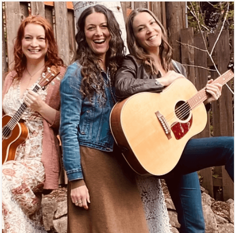 Three smiling women posing outdoors with a ukulele and an acoustic guitar in Central Oregon.