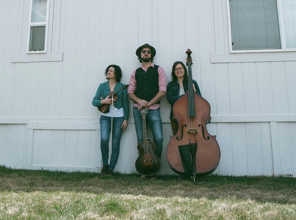 Three musicians standing against a white wall holding a violin, guitar, and upright bass in Central Oregon.
