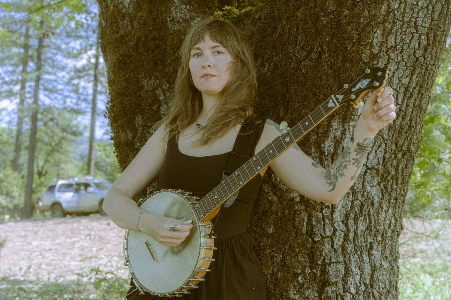 A woman with tattoos holding a banjo while leaning against a large tree in Central Oregon.