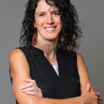 A woman with curly dark hair smiles with her arms crossed against a grey background in Central Oregon.