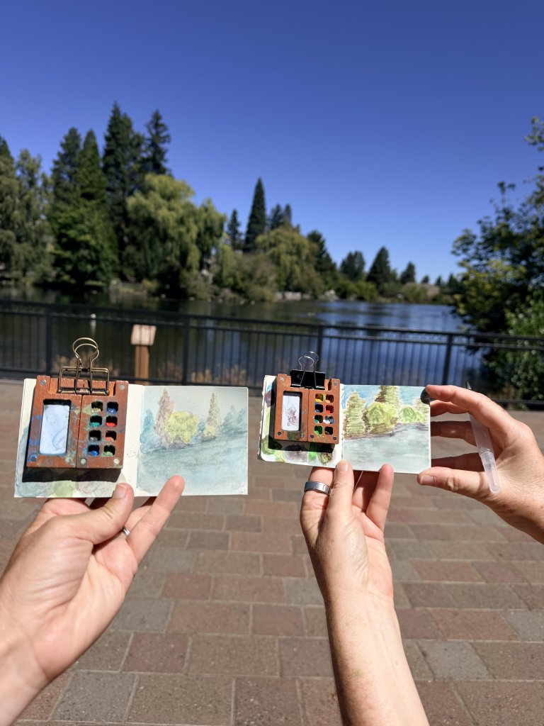 Two hands hold small watercolor sketchbooks featuring paintings of a lake and trees in Central Oregon.