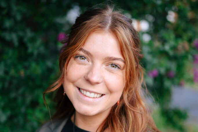 A smiling young woman with auburn hair poses outdoors against a leafy green background in Central Oregon.