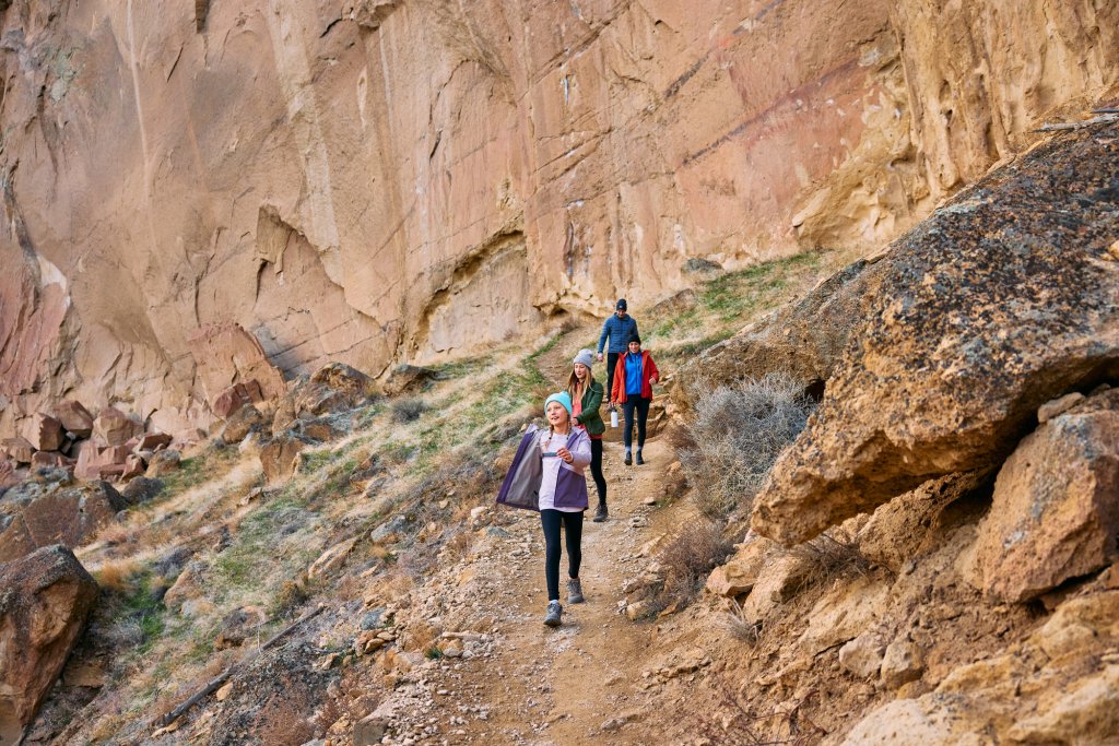 A group of people hiking on a narrow dirt trail beside a large rock wall in Central Oregon.