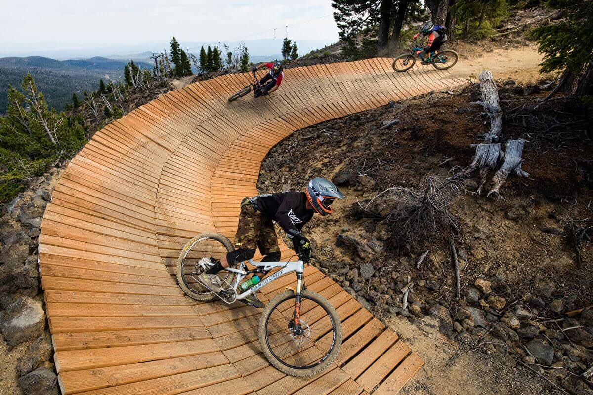 Three mountain bikers riding on a curved wooden trail feature on a mountainside in Central Oregon.