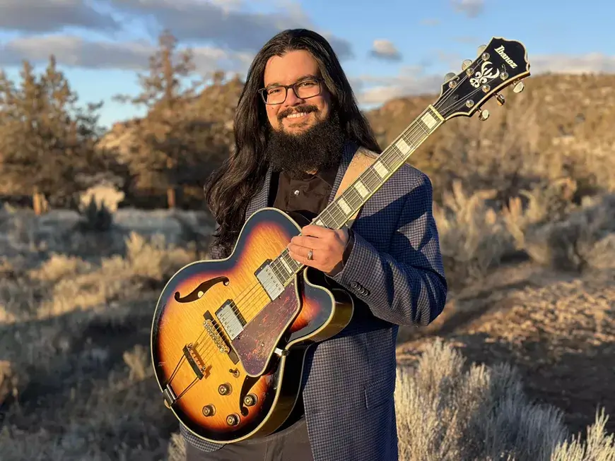 A smiling man with long hair and a beard holding an Ibanez hollow-body guitar in Central Oregon.