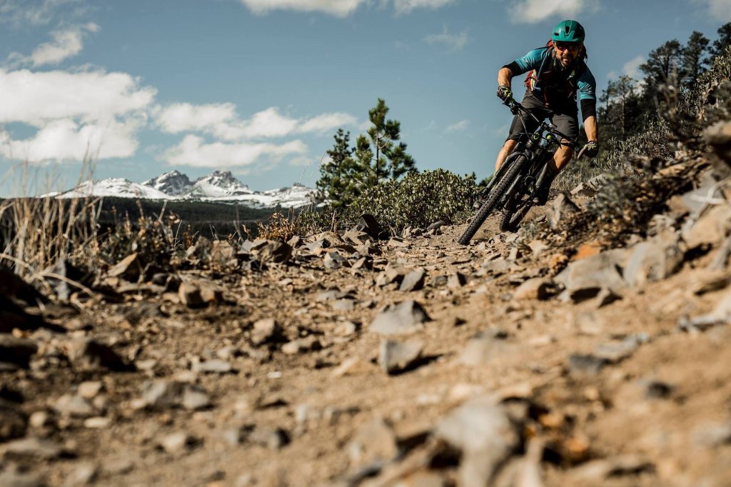 A mountain biker rides down a rocky trail with snow-capped mountains in the background in Central Oregon.