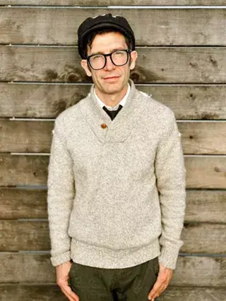 A man wearing a cap, glasses, and a light-colored sweater against a wooden background in Central Oregon.