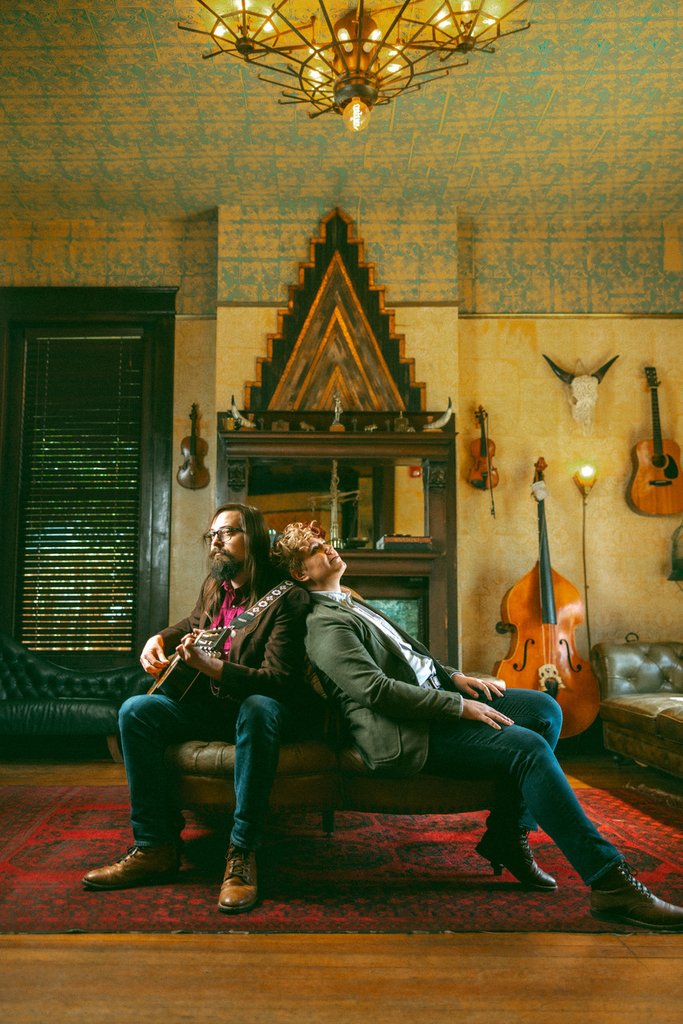 Two musicians sit back-to-back on a leather couch in a vintage-styled room in Central Oregon.
