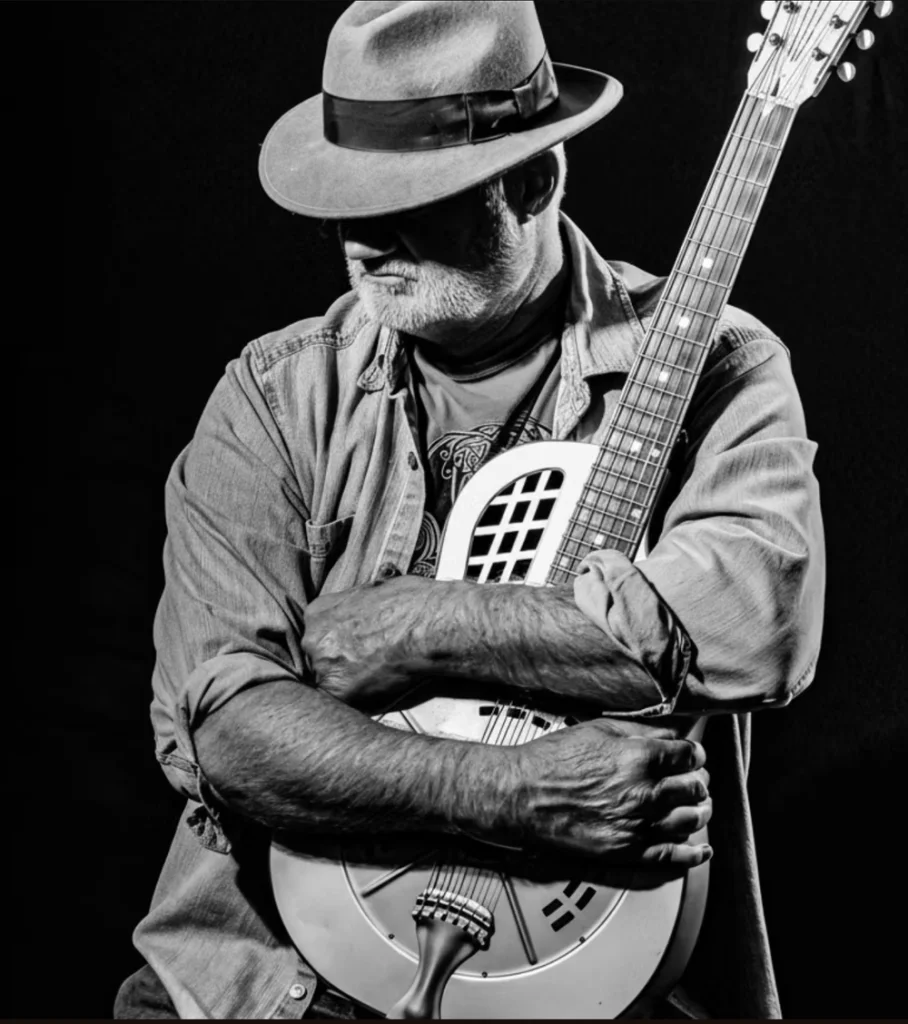 A bearded man in a fedora holding a resonator guitar against his chest in Central Oregon.