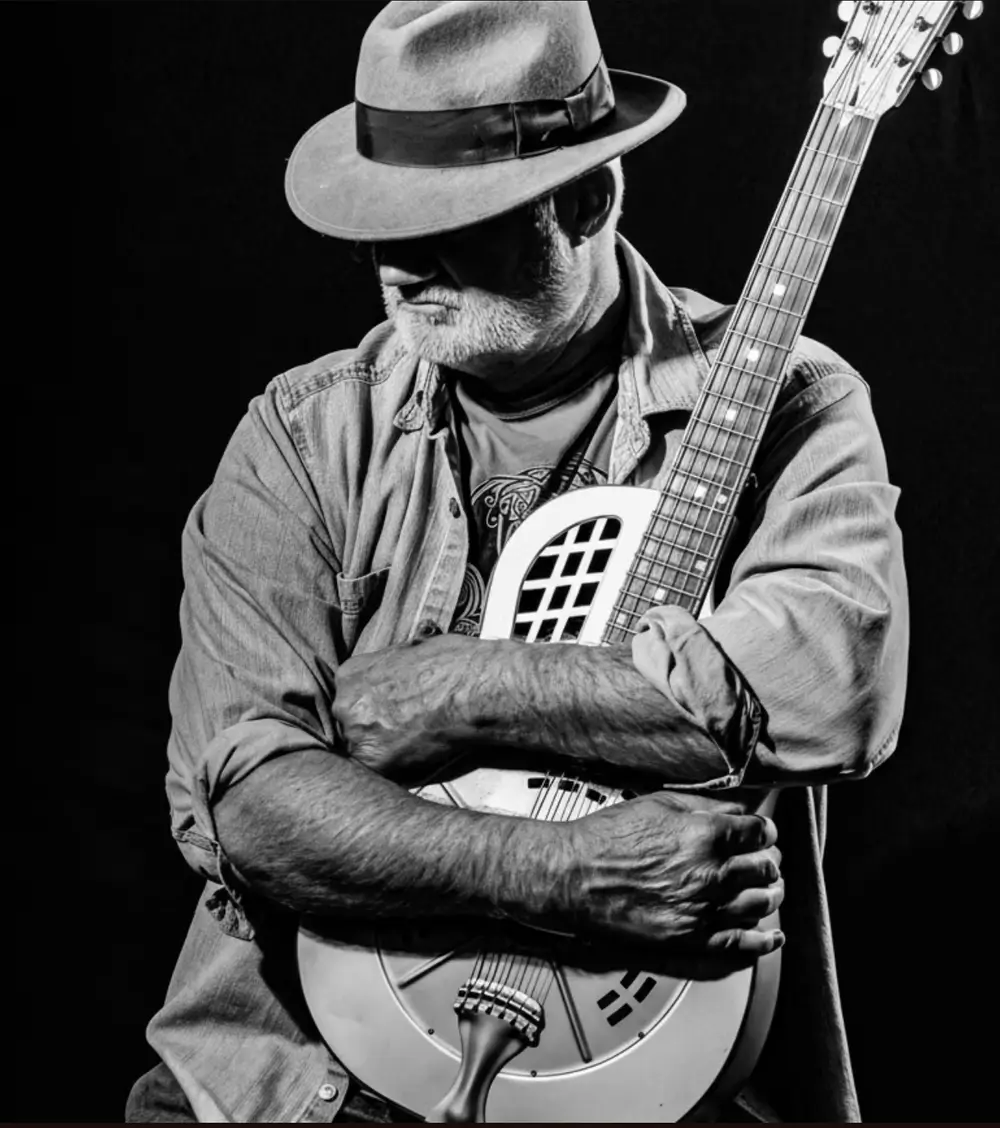 A bearded man in a fedora holding a resonator guitar against his chest in Central Oregon.