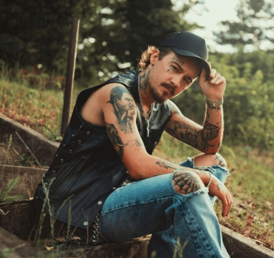 A tattooed man in a leather vest and cap sits on concrete stairs outdoors in Central Oregon.