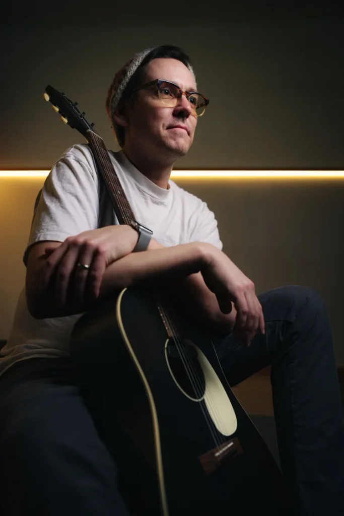 A man wearing glasses and a beanie poses with an acoustic guitar in Central Oregon.