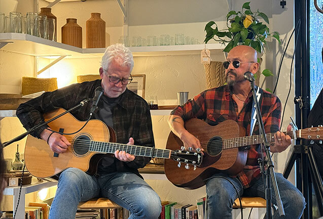 Two men playing acoustic guitars while sitting on stools in a shop in Central Oregon.