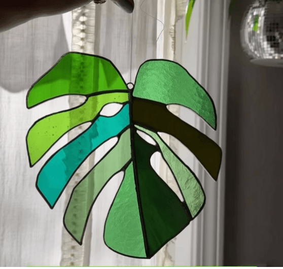 A stained glass monstera leaf suncatcher hanging in front of a window in Central Oregon.