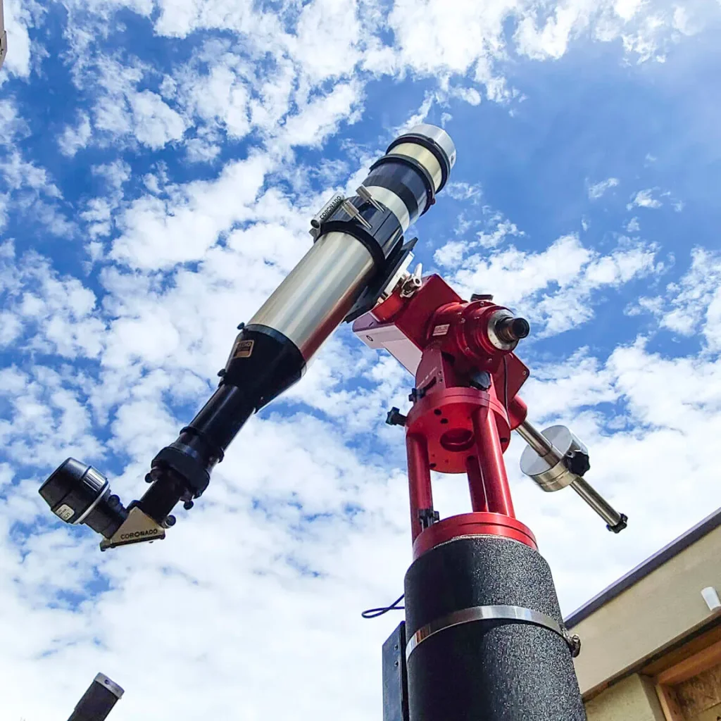 A solar telescope mounted on a red equatorial mount against a cloudy blue sky in Central Oregon.