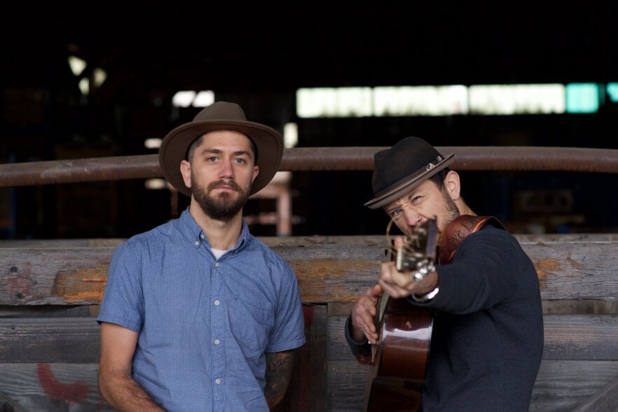Two men wearing hats, one holding a guitar, standing in front of a wooden fence in Central Oregon.