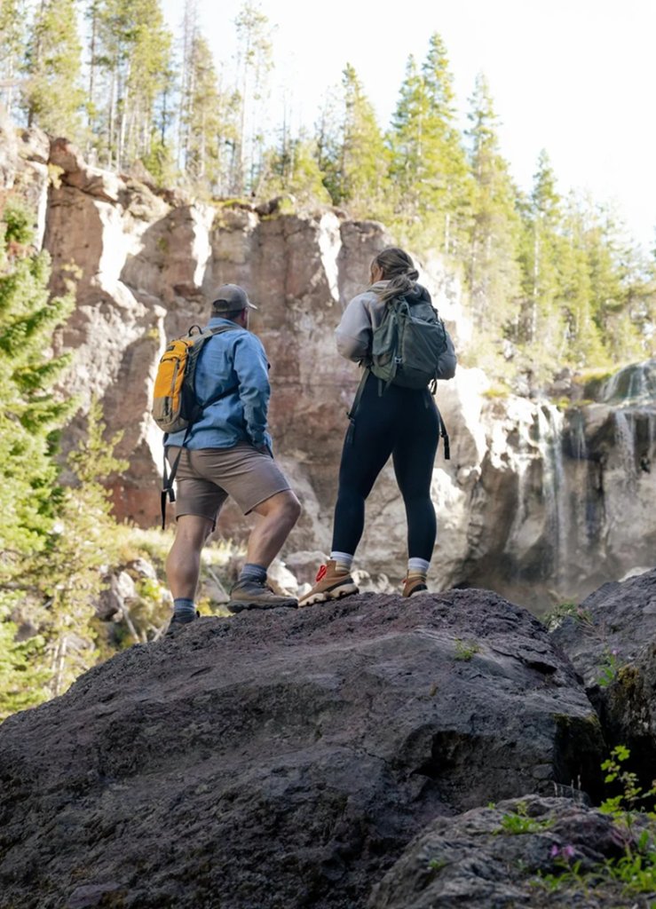 A couple with backpacks stands on a large rock overlooking a waterfall in Central Oregon.