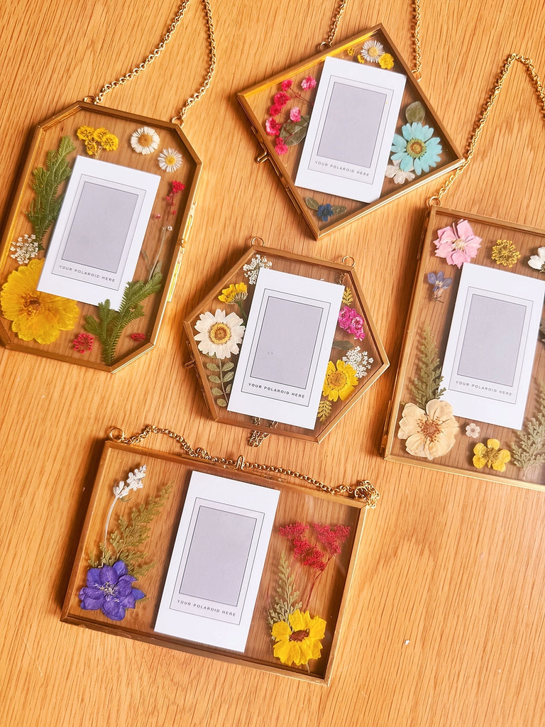 Five hanging glass frames containing pressed flowers and blank polaroid-style inserts on wood in Central Oregon.