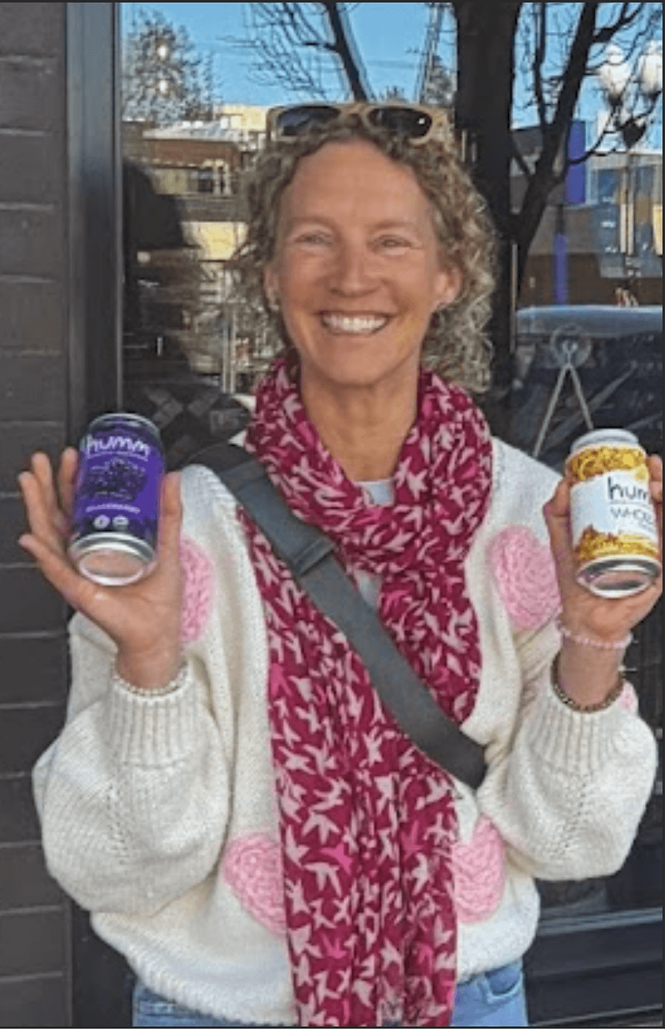 A smiling woman with curly hair holds two cans of Humm kombucha in Central Oregon.