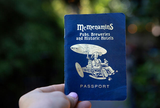 A hand holding a blue McMenamins passport booklet against a blurred outdoor background in Central Oregon.
