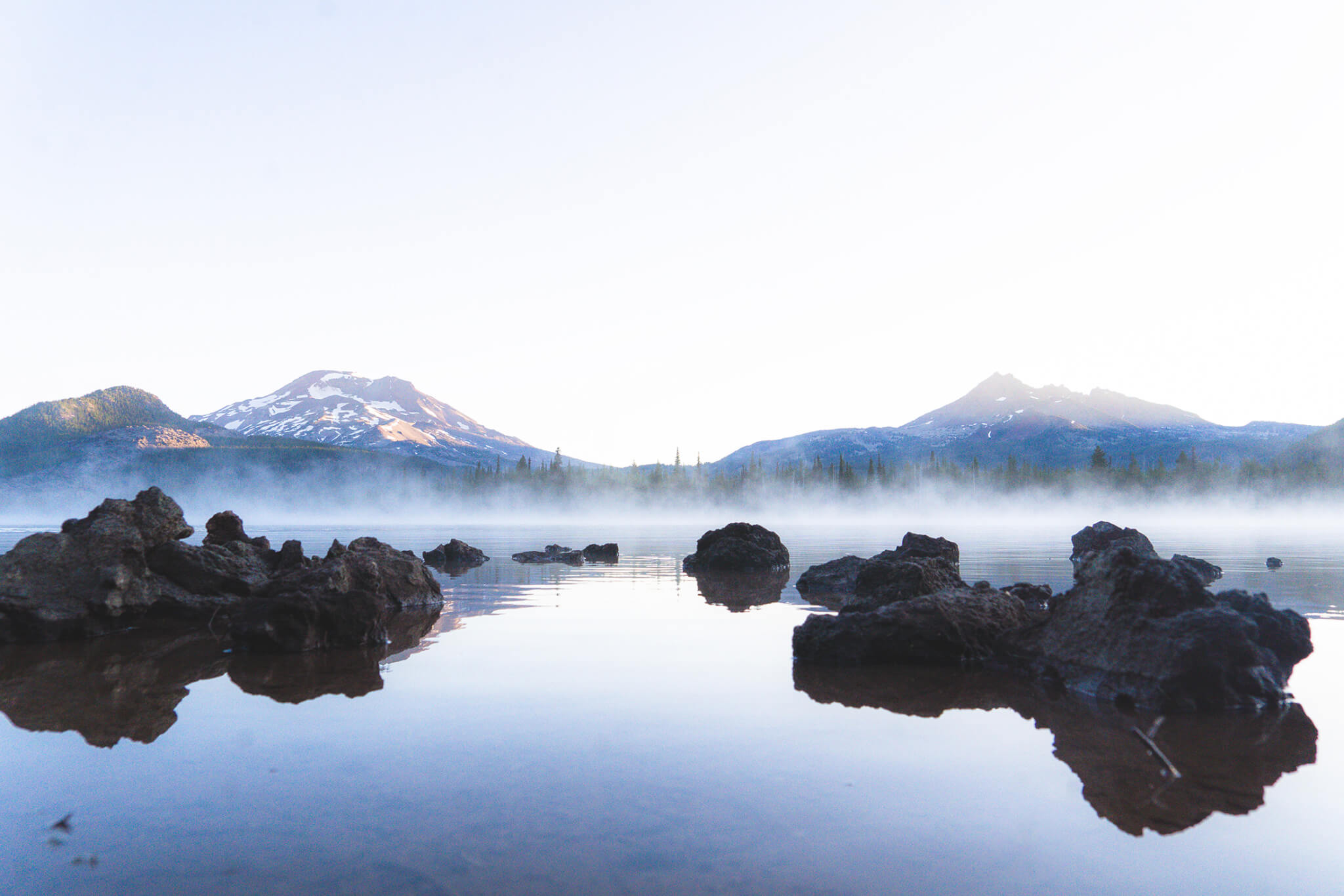 Dark volcanic rocks sit in a misty lake with snow-capped mountains in the background in Central Oregon.