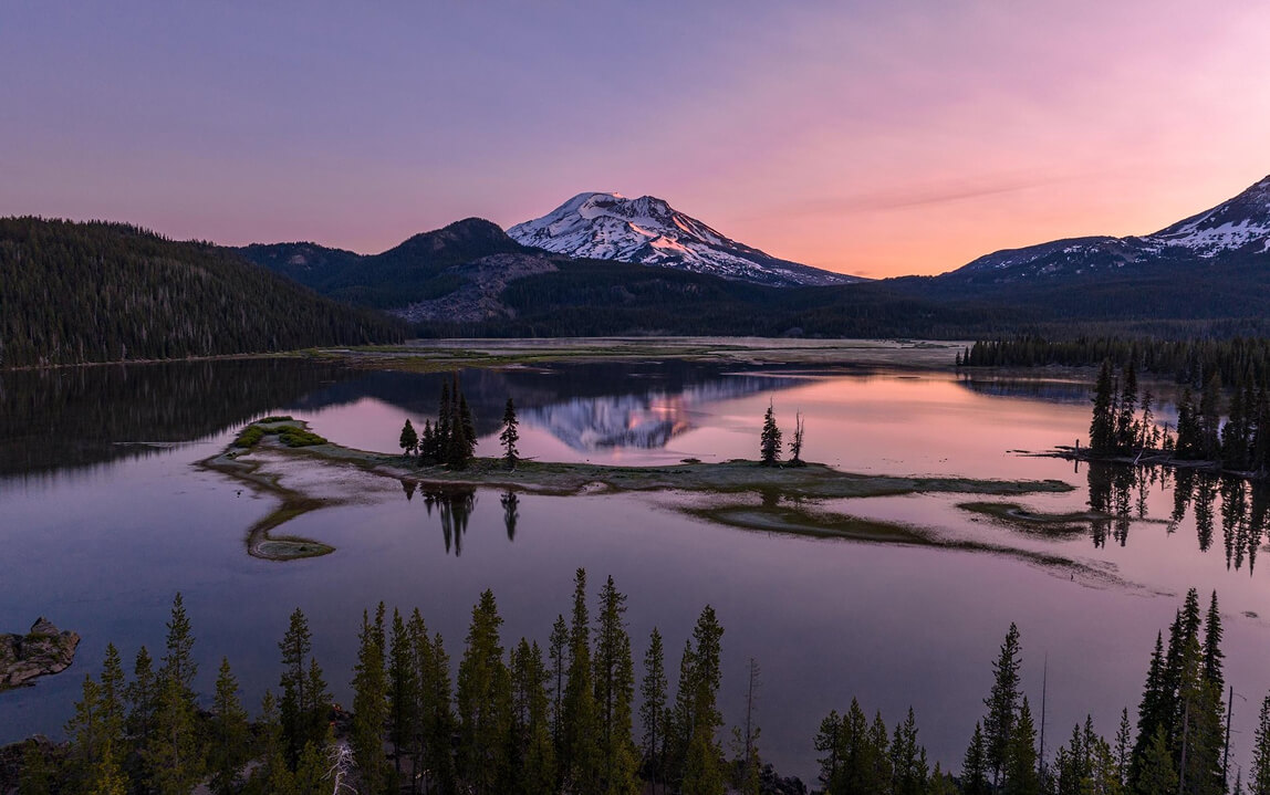 A snow-capped mountain reflects in a calm lake during a pink sunset in Central Oregon.