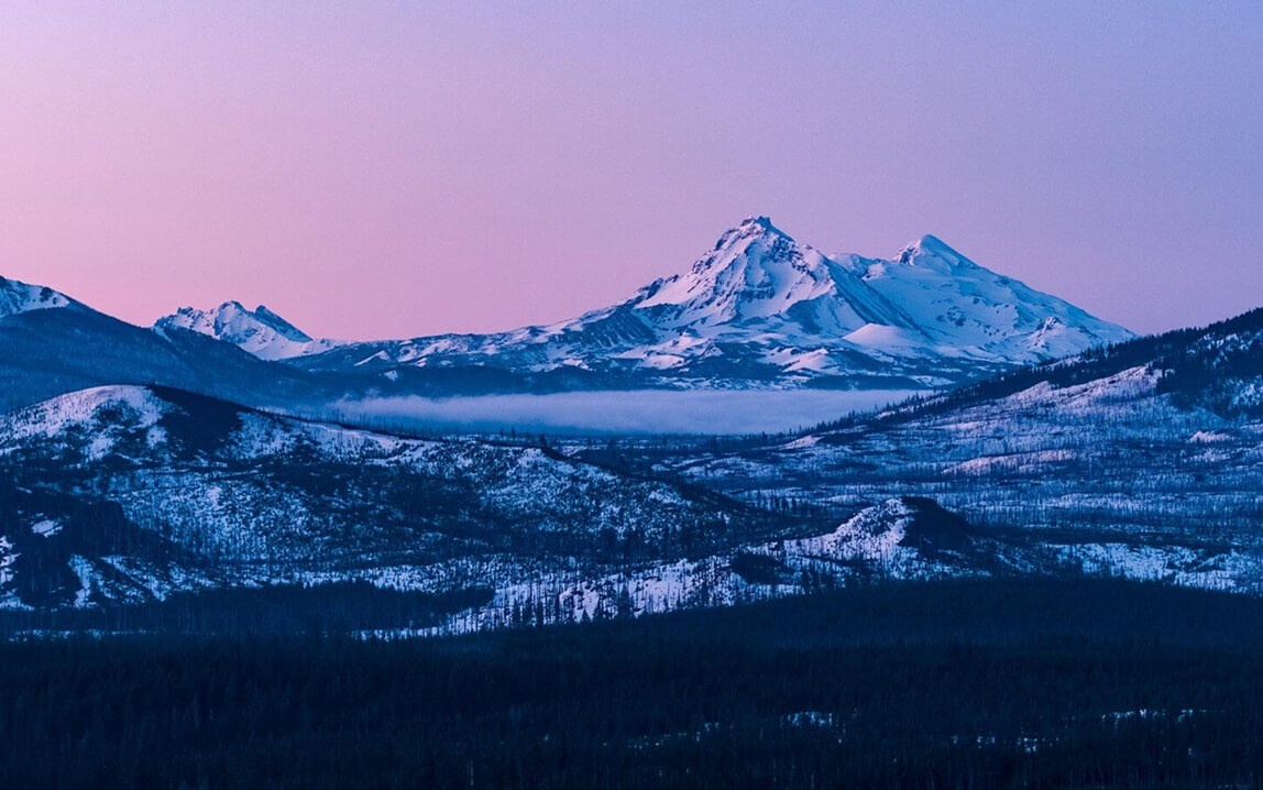 Snow-capped mountains rise above a misty valley under a soft purple sunset sky in Central Oregon.