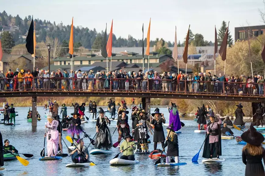 People in witch costumes paddleboard on a river under a crowded pedestrian bridge in Central Oregon.
