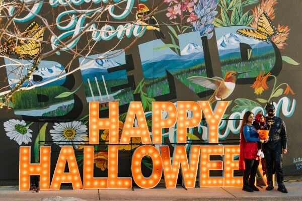 A family in superhero costumes posing in front of a Happy Halloween light display in Central Oregon.