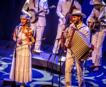 A band in white outfits performs on stage with a violinist and accordion player in Central Oregon.