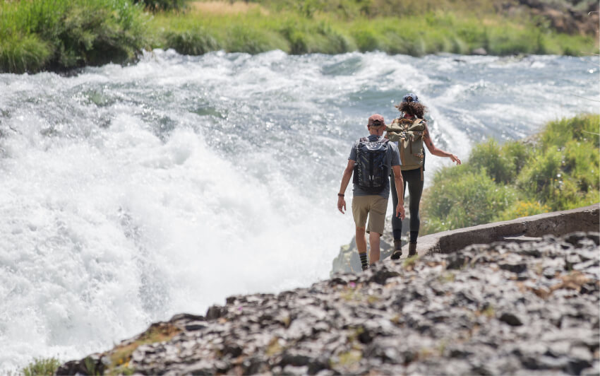 Two hikers with backpacks walk along a path beside a rushing river in Central Oregon.