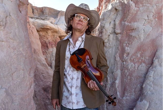 A man in a cowboy hat and blazer holds a violin in a desert canyon in Central Oregon.