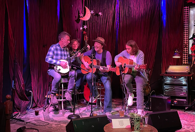 A four-piece folk band performing on stage with acoustic instruments and microphones in Central Oregon.