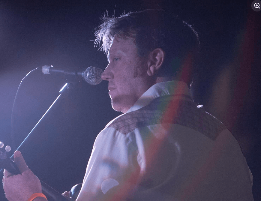 A man in a button-down shirt performing with a guitar and microphone on stage in Central Oregon.