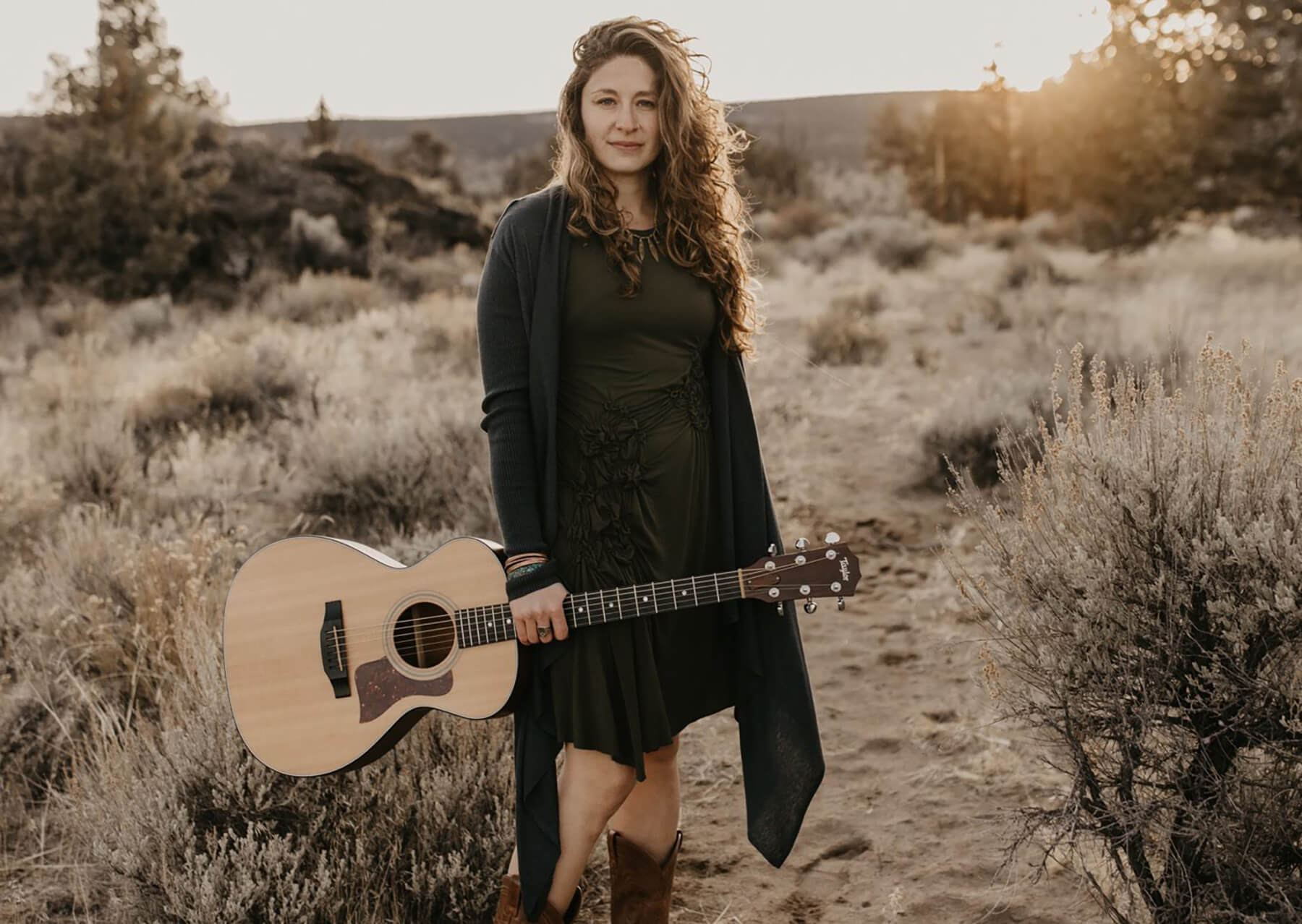 A woman with long wavy hair holding an acoustic guitar in a desert landscape in Central Oregon.