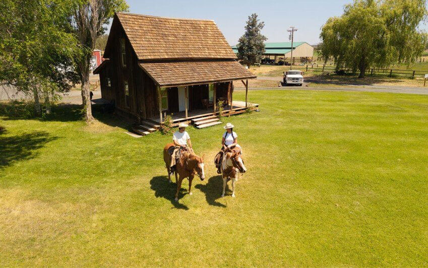 Two people on horseback ride across a grassy field in front of a rustic cabin in Central Oregon.