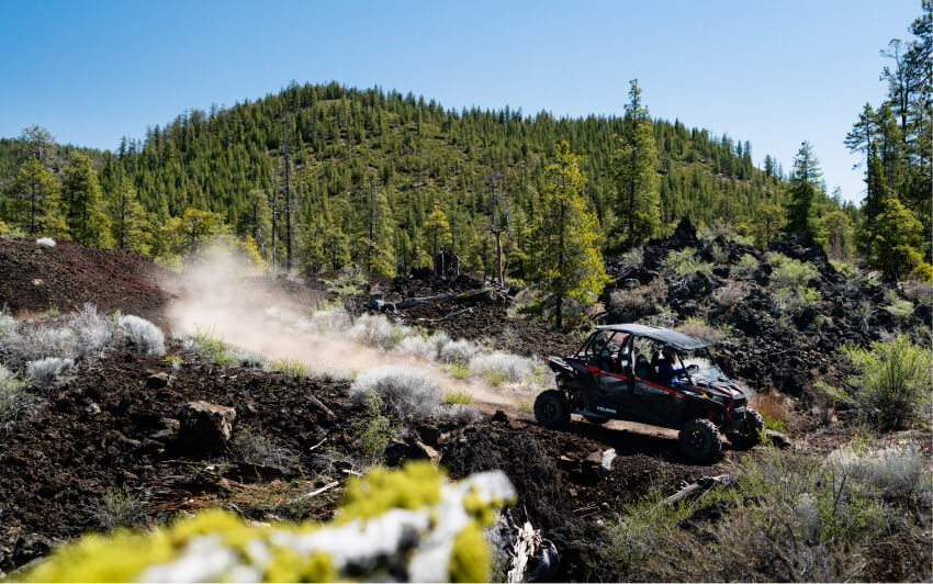 A side-by-side vehicle driving on a dusty trail through a volcanic landscape in Central Oregon.