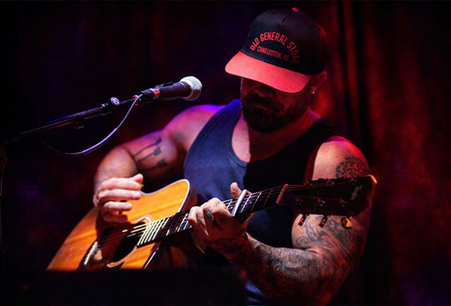 A tattooed man wearing a cap plays an acoustic guitar on stage in Central Oregon.