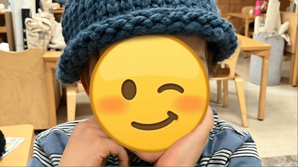 A child wearing a blue knitted hat with their face covered by a winking emoji in Central Oregon.