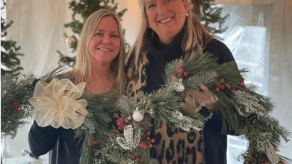 Two smiling women holding a decorated Christmas garland with a large bow in Central Oregon.