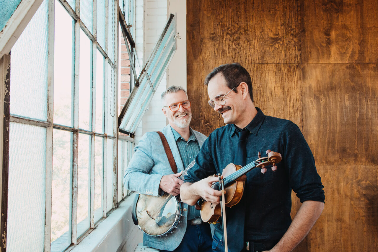Two men smiling while playing a banjo and a violin near a window in Central Oregon.