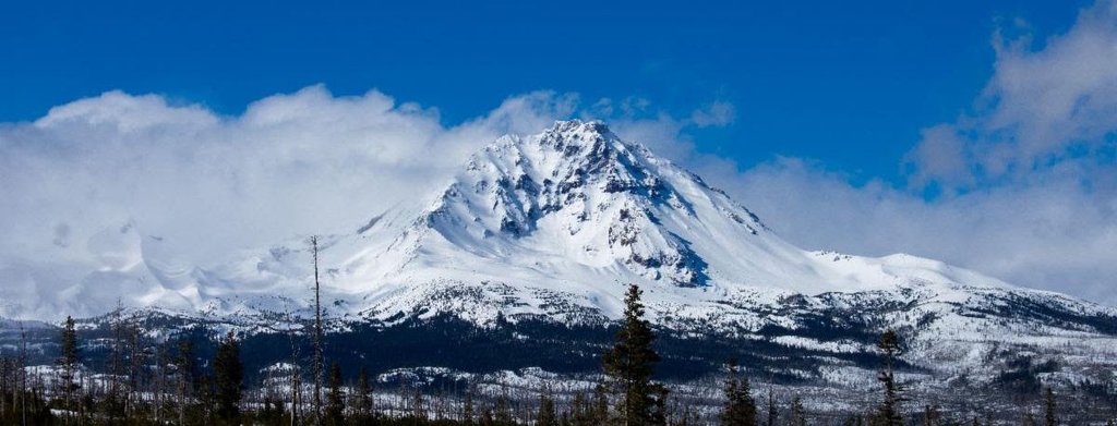 A majestic snow-covered mountain peak rises against a bright blue sky with scattered clouds in Central Oregon.
